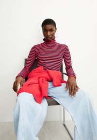 Gestreiftes Turtleneck-Oberteil in Rot und Marineblau, kombiniert mit lockeren hellblauen Jeans, über die die rote Strickjacke gelegt ist.