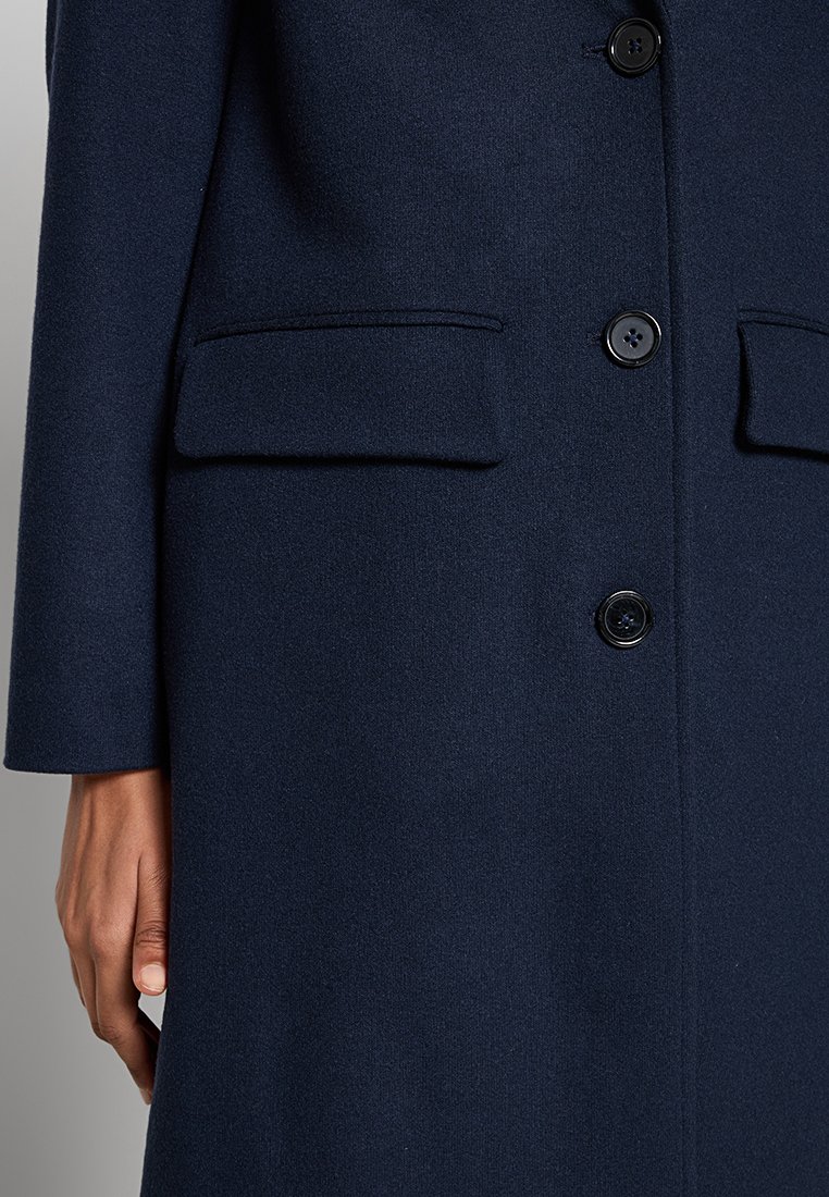 Cappotto di lana blu navy con chiusura frontale a bottoni, due tasche laterali e maniche lunghe. Tessuto liscio con vestibilità sartoriale.
