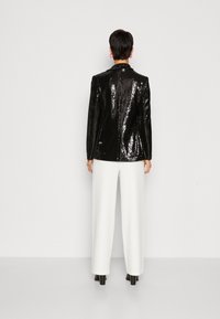 Černý lesklý blejzr s přiléhavým střihem, dlouhými rukávy a klopami, zkombinovaný s bílými kalhotami s vysokým pasem a černými podpatky.