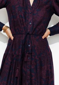 Robe à manches longues et à boutons en bleu marine foncé avec un motif floral bordeaux. Présente une taille nouée et un ourlet texturé avec des accents de pompons.