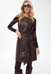 Blondynka w dużych okularach przeciwsłonecznych, przezroczystej czarnej bluzce w kwiaty z długim rękawem, bezrękawnikowej brązowej skórzanej sukience z paskiem oraz ciemnych spodniach i butach.