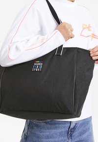 Osoba trzymająca dużą czarną torbę na ramię z logo "Puma Tailored for Sport", ubrana w białą koszulę z długim rękawem i niebieskie dżinsy.