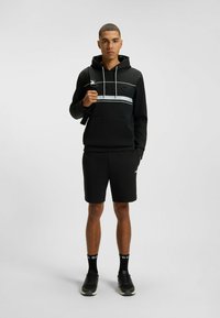 Felpa nera con cappuccio e logo in rilievo, abbinata a pantaloni corti neri e sneakers nere. Design semplice e sportivo.