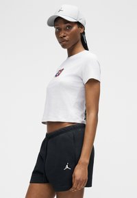 Γυναίκα με λευκό καπέλο, λευκό κοντό μπλουζάκι και μαύρο σορτς με αθλητικά λογότυπα, στέκεται μπροστά σε απλό φωτεινό φόντο.