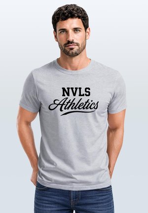 Mann mit dunklem lockigem Haar und Bart, der ein hellgraues T-Shirt mit schwarzem Text "NVLS Athletics" trägt, steht vor einem schlichten hellen Hintergrund.