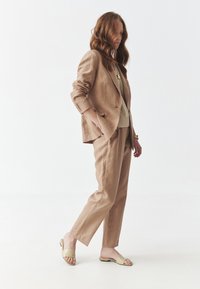 Costume en lin beige avec veste cintrée et pantalons droit, associé à des sandales à glissière de couleur claire ; texture lisse et coupe décontractée.