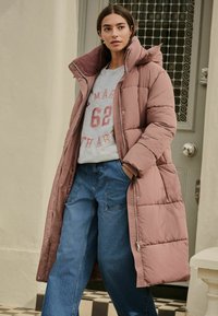 Geacă pufos roz deschis cu guler înalt, asociată cu un hanorac gri cu text roșu și pantaloni denim albaștri largi. Tesătura are un aspect texturat, iar cusăturile sunt vizibile.