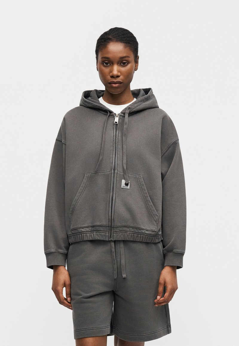 Donna che indossa una felpa con cappuccio grigio scuro con zip e tasche frontali e pantaloncini abbinati, in piedi davanti a uno sfondo chiaro e uniforme.