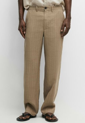 Mand iført beige pinstripe viddebukser og sorte sandaler står foran en ensfarvet baggrund.