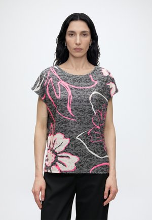 Femeie cu păr lung, ondulat și închis la culoare, purtând un top gri cu mâneci scurte cu imprimeu floral mare roz și alb și pantaloni negri, stând în picioare pe un fundal alb.