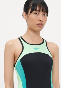 Maillot de bain noir et vert menthe avec un col haut et des fines bretelles, arborant un logo Speedo et un design en color block contrasté.
