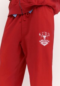 Czerwone spodnie dresowe z troczkiem w pasie i białym nadrukowanym emblematem z napisem "STWD Forever Authentic 1991" na górnej części uda.
