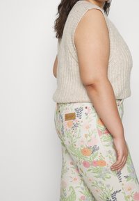 Top sin mangas de punto beige combinado con jeans de color claro con estampado floral y un parche de marca marrón en el bolsillo trasero.