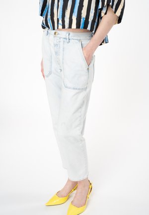 Osoba nosząca jasnoniebieskie jeansy z wysokim stanem, paskowany niebiesko-beżowy top oraz jasne żółte buty typu slingback z ostrym noskiem, stojąca z jedną ręką w kieszeni.