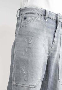 Primer plano de unos jeans de mezclilla gris claro con parches desgastados y costuras visibles en la parte superior del muslo y la cadera.