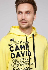 Sudadera con capucha amarilla con texto impreso en negro y adornos. Incluye un cordón de rayas en blanco y negro y una tela texturizada.