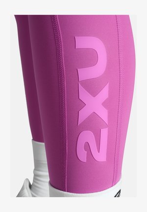 Leggings sportivi rosa con texture liscia, caratterizzati da un grande logo bianco "2XU" sulla coscia, cuciture a contrasto e un polsino bianco alla caviglia.
