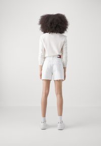 Sudadera blanca corta y pantalones cortos de mezclilla blanca deshilachados. Zapatillas con acentos metálicos y calcetines blancos. El cabello es voluminoso y rizado.