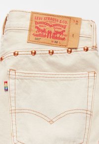 Jeans de mezclilla blanca con remaches de cobre, costuras naranjas y una etiqueta roja en la cinturilla. Tiene una pequeña etiqueta de arcoíris en el bolsillo.