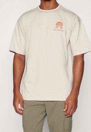 Hombre que lleva una camiseta beige de Columbia con logo naranja en el pecho y pantalones verde oliva, de pie frente a un fondo blanco liso.