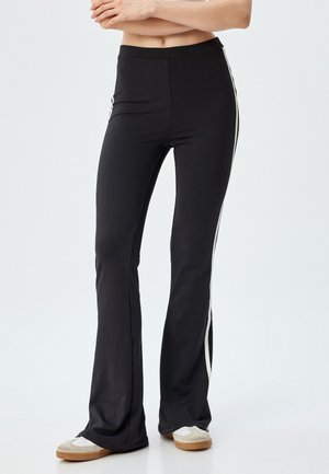 Pantalones de campana negros con una textura suave, con franjas laterales blancas. Cintura elástica para un ajuste ceñido. Los zapatos son beige con acentos blancos.