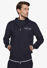 Marineblå zip-up sweatshirt i strikstof med to forlommer, justerbar snor i hætten og kontrasterende hvide ribbede manchetter og kant.