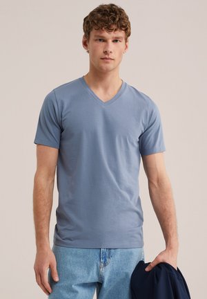 Jauns vīrietis ar cirtainiem matiem, valkājot gaiši zilu V-veida kakla T-kreklu un gaiši zilas džinsas, labajā rokā tur tumšu jaku.