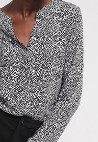 Zwart-witte blouse met een klein, verspreid loofpatroon, v-hals en knoopdetails aan de voorkant. Zachte stof met lange mouwen.