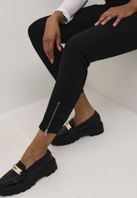 Pantaloni neri aderenti con cerniera alla caviglia, abbinati a mocassini neri con un accento dorato. Texture liscia e design su misura.