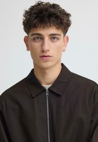 Veste marron à fermeture éclair avec col, en tissu texturé, et une fermeture éclair à l'avant. Le modèle a les cheveux bouclés et les yeux bleus.