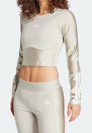 Weiblicher Torso mit beigem, langärmligem Cropped-Sporttop und passenden Leggings mit hohem Bund, versehen mit weißen Adidas-Logos und Streifen.