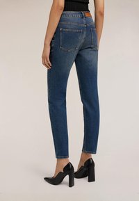 Žena v modrých džínech s vysokým pasem a černých botách na blokovém podpatku, zobrazena zezadu na jednobarevném pozadí.