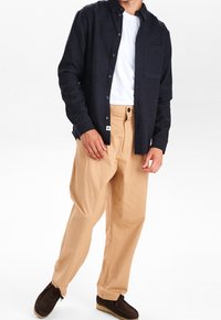 Granatowa koszula z guzikami nałożona na białą koszulkę, w połączeniu z luznymi beżowymi spodniami i brązowymi butami wsuwanymi. Posiada kieszeń na piersi i guziki.