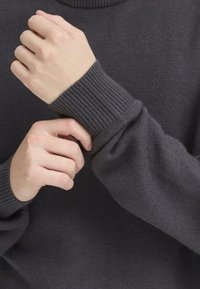 Grijze gebreide trui met ribbenmanchetten, met een gladde textuur en een hoge halslijn, waarop handen te zien zijn die de mouw aanpassen.