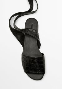 Schwarze Slidesandalen aus strukturiertem Kunst-Krokodilleder. Mit einem breiten Riemen über dem Fuß und einem dünnen, geschwungenen Knöchelriemen.