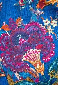 Broderie florale lumineuse en rouge, violet, jaune et blanc sur un fond de tissu bleu texturé avec des motifs détaillés de feuilles et de fleurs.