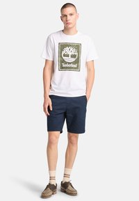 Camiseta blanca con el logo verde de Timberland, pantalones cortos azul marino, calcetas beige con rayas marrones y zapatos marrones. El modelo está de pie con las manos en los bolsillos.