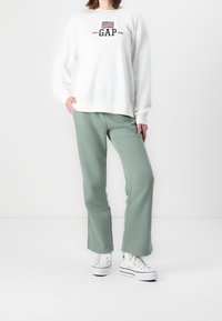 Personne portant un sweat à capuche blanc GAP avec un drapeau américain, un pantalon de jogging vert sauge et des baskets montantes blanches sur un fond blanc uni.