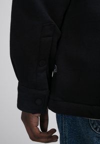 Zwarte sweatshirt met een gestructureerde afwerking, voorzien van knoopmanchetten en een klein merklabel aan de zijkant. Spijkerbroek zichtbaar daaronder.