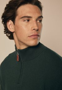 Maglione zip-up verde scuro con colletto alto a coste. Presenta una zip argentata e un design in maglia a trama.