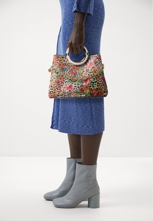 Vrouw draagt een blauwe geribbelde gebreide jurk en grijze enkellaarsjes, houdt een tas met bloemen- en luipaardprint met ronde handvatten en een gouden ketting vast.