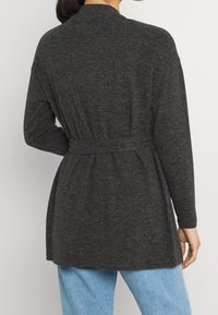 Mörkgrå, texturerad kofta med bälte i midjan och långa ärmar, försedda med en sjalkrage. Bärs över ljusblå jeans.