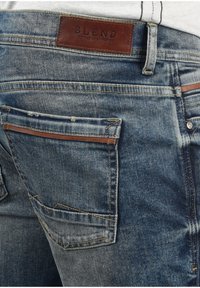 Jeans de mezclilla azul con acabado desgastado; presenta un parche de cuero marrón con la marca "BLEND" y dos bolsillos traseros con costuras en contraste.