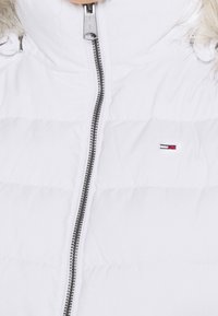 Biała kurtka puchowa z odpinanym futrzanym kapturem, z srebrnym zamkiem błyskawicznym, pikowanym wzorem i małym logo po lewej stronie.