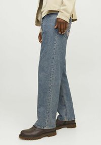 Sinise denim teksad, millel on lai istuvus, detailse tagatasku disainiga ja neid kantakse pruuni kingadega. Kudel on kerge tekstuur.