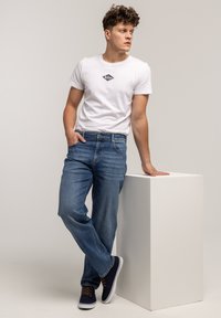 Biała bawełniana koszulka z małym ciemnym logo, w połączeniu z niebieskimi jeansami. Model ma na nogach granatowe trampki i stoi swobodnie oparty o biały postument.