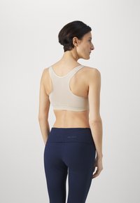 Sujetador deportivo beige con diseño de espalda nadadora, hecho de tela elástica, presenta costuras planas. Combinado con leggings de talle alto en color azul marino que exhiben un logo.