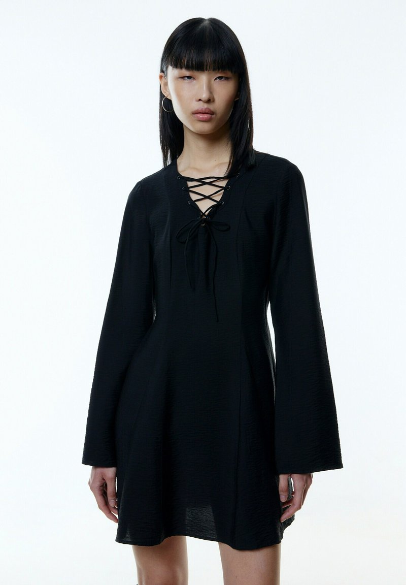 Schwarzes Kleid mit langen Glockenärmeln, einem Schnürkragen und einem strukturierten Stoff, der von der Taille bis zum Saum aufflackert.