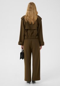 Bruine jumpsuit met een taillering, brede pijpen en een gelaagde kraag. Model houdt een kleine zwarte gestructureerde handtas vast. Gladde stofafwerking.