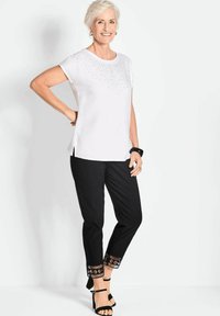 Witte kortarmige top met strass-detail, gecombineerd met zwarte broek met een patroonrand en zwarte sandalen met bandjes.